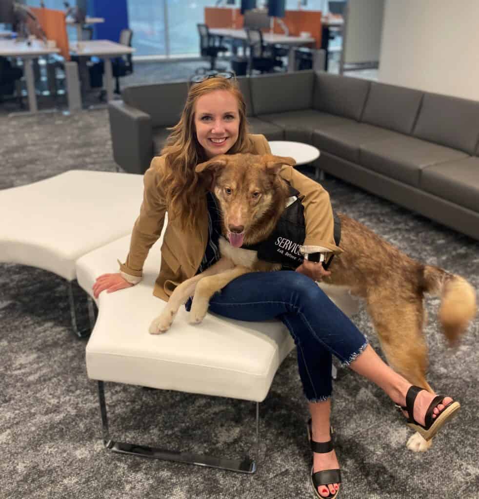 Kaydin Downey and her Service Dog and medical alert dog Hamilton provide consulting services at offices. Image shows Kaydin in an office, seated on a white leather sofa. Hamilton is applying pressure on her lap with his paws and torso.